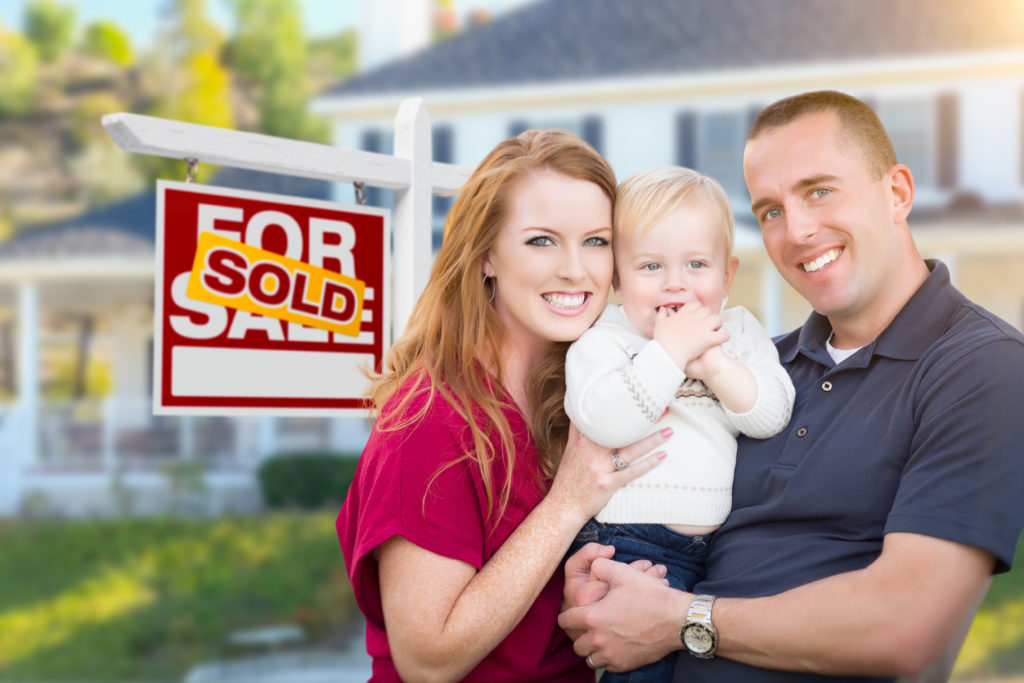 Happy Young Military Family in Front of Sold For Sale Real Estate Sign and New House purchased through Goldfarb Capital.